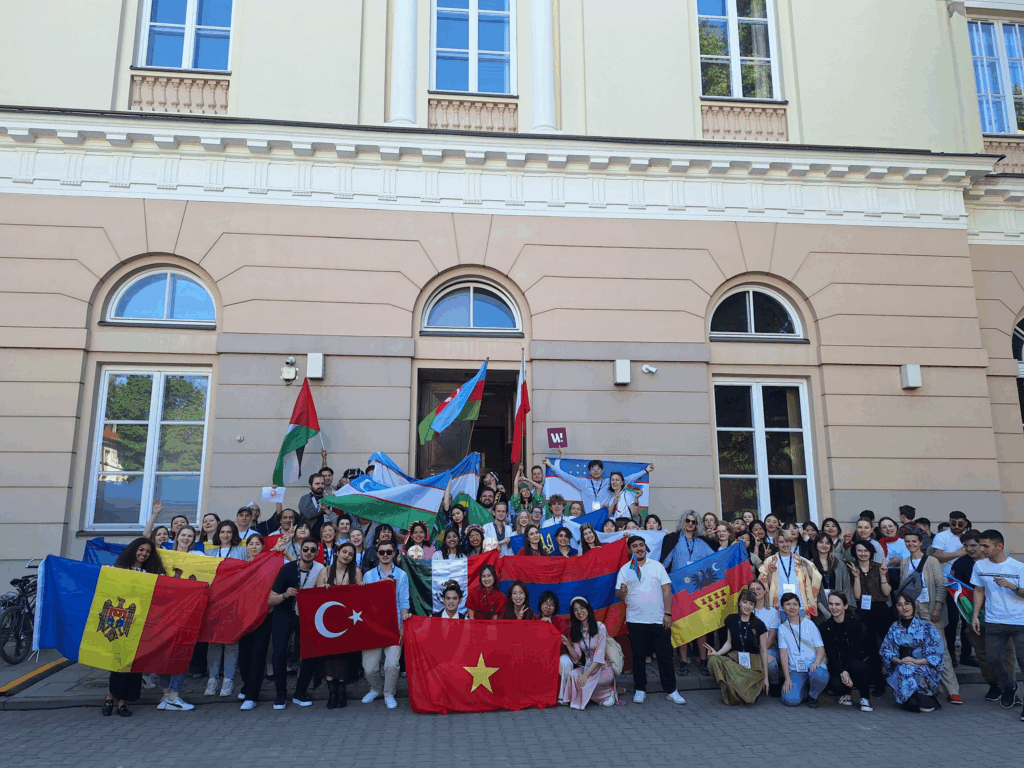 The image shows a group of people from various countries posing for a photo. Some of them are holding their country's flag.